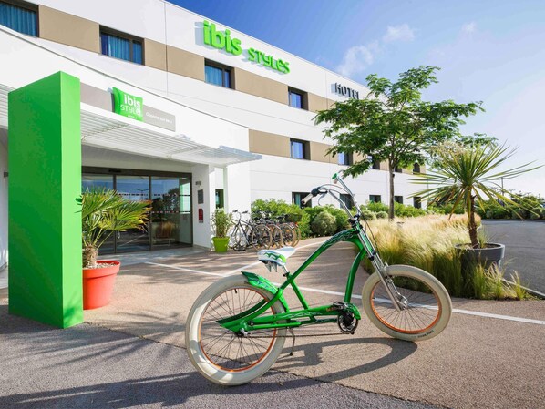 Un hotel llamado 'ibis Styles' con un diseño moderno, una bicicleta estacionada adelante y plantas en macetas alrededor.
