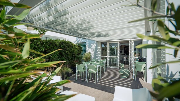 Une terrasse extérieure avec des tables et des chaises blanches, une entrée avec une porte vitrée et un espace abrité doté d’un toit en métal.
