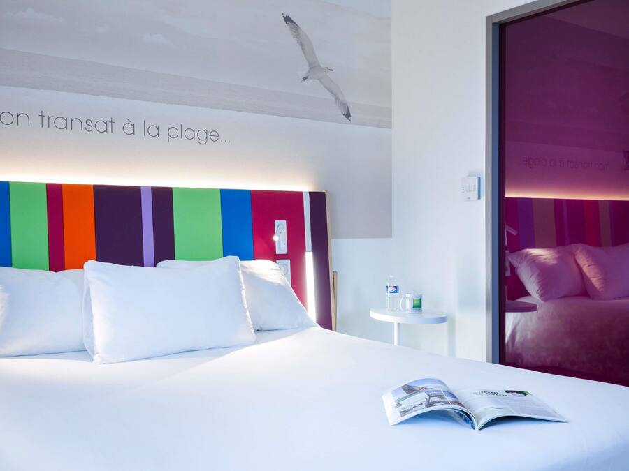Habitación de hotel con una cama, una revista sobre la cama, una mesita con una botella y una taza, y un cabecero a rayas de colores.