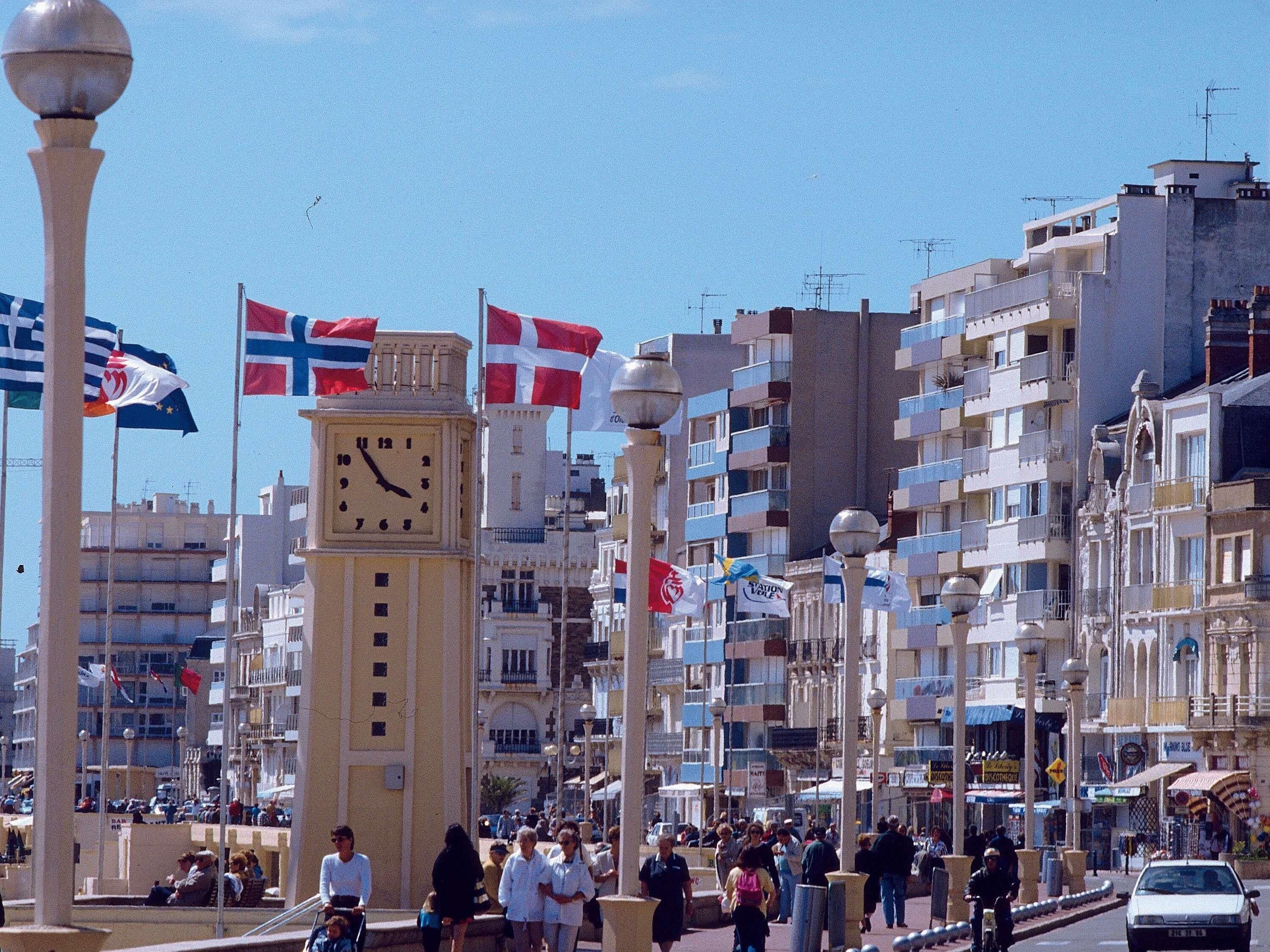 Une rue animée en plein centre-ville, avec un beffroi imposant, divers drapeaux et des immeubles à plusieurs étages.