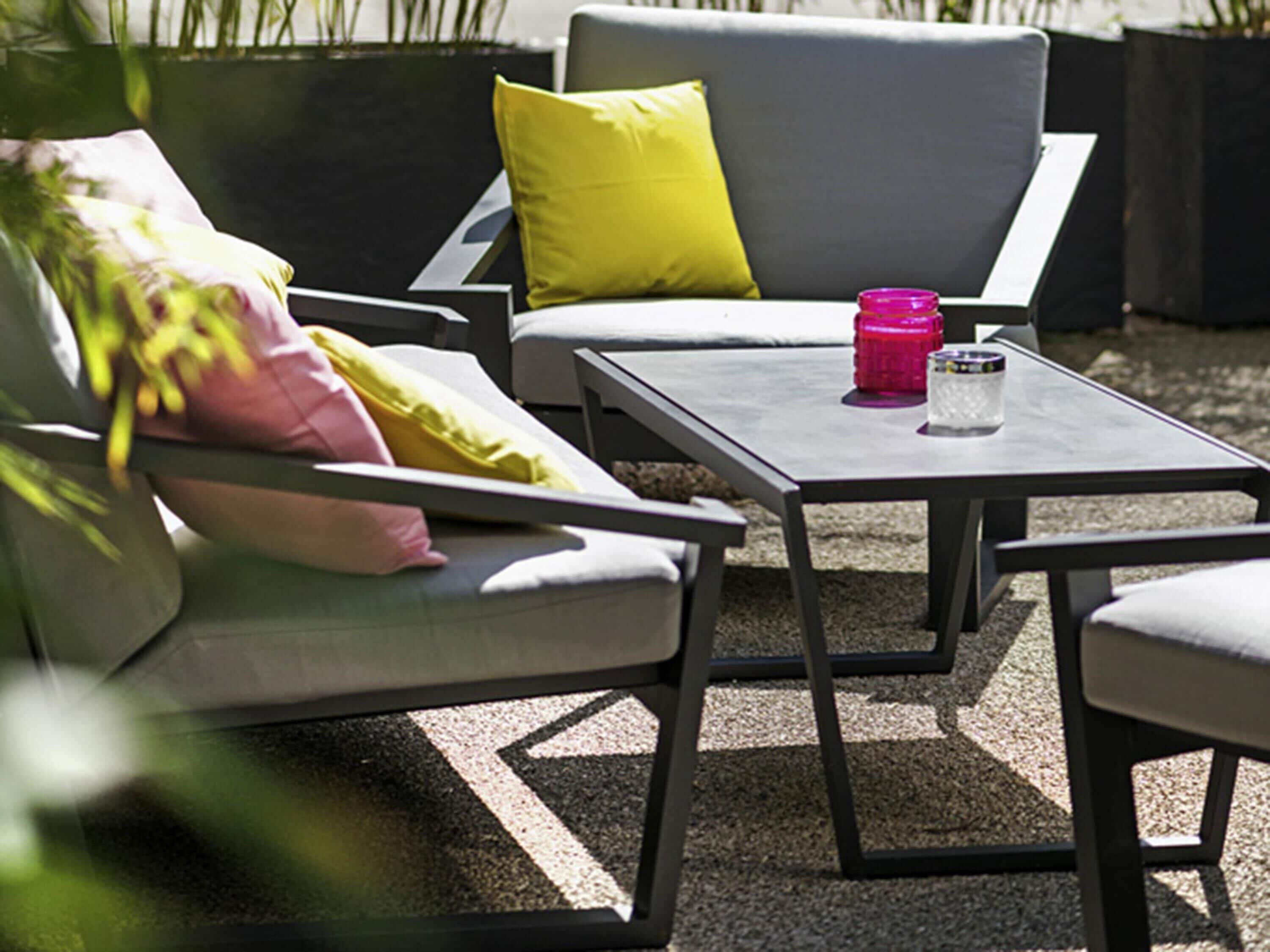 An outdoor seating area with a gray sofa, yellow and pink cushions, and a small table with a pink container and a glass.