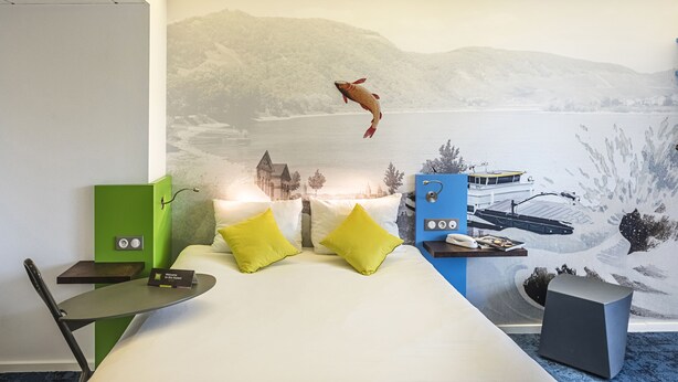A hotel room with a bed, two yellow pillows, a green side table, a blue side table, and a mural of a fish jumping out of water. - ibis Styles Strasbourg Avenue du Rhin (Strasbourg)