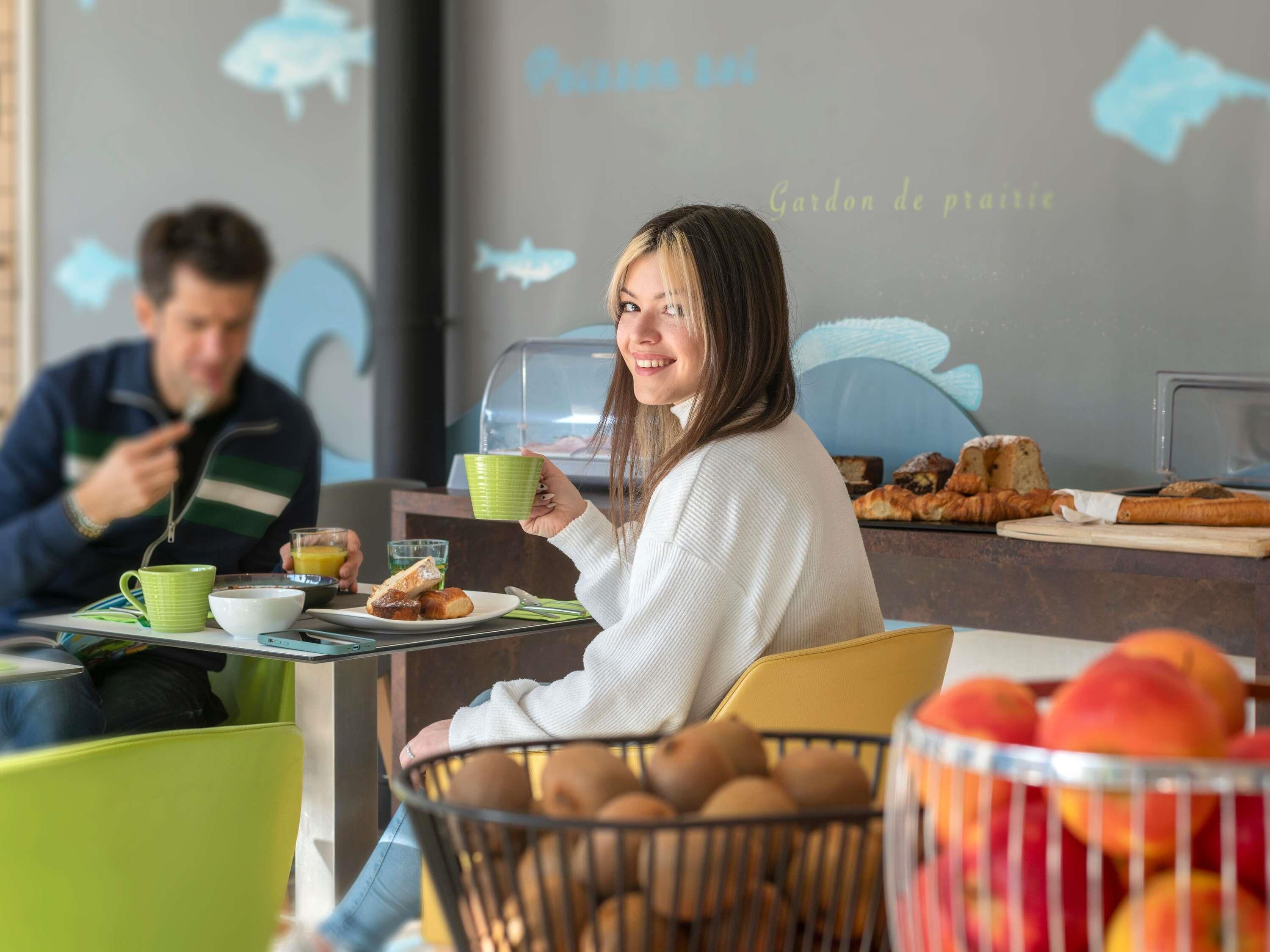 A breakfast setting with a person holding a green cup, another person eating, and a variety of food items on the table.