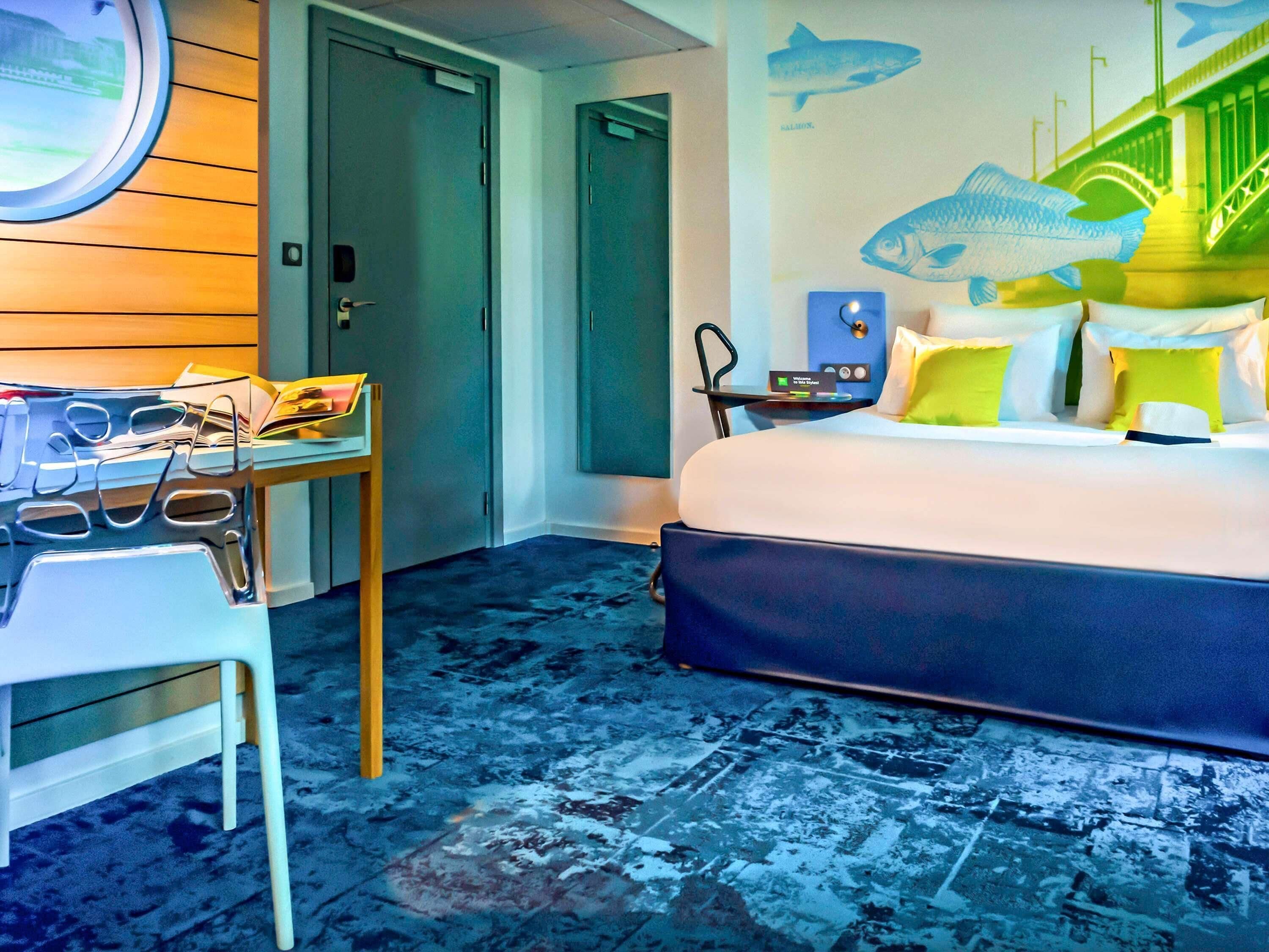 Ett hotellrum med en säng, ett skrivbord med en bärbar dator, en stol och en vägg med illustrationer av fiskar.