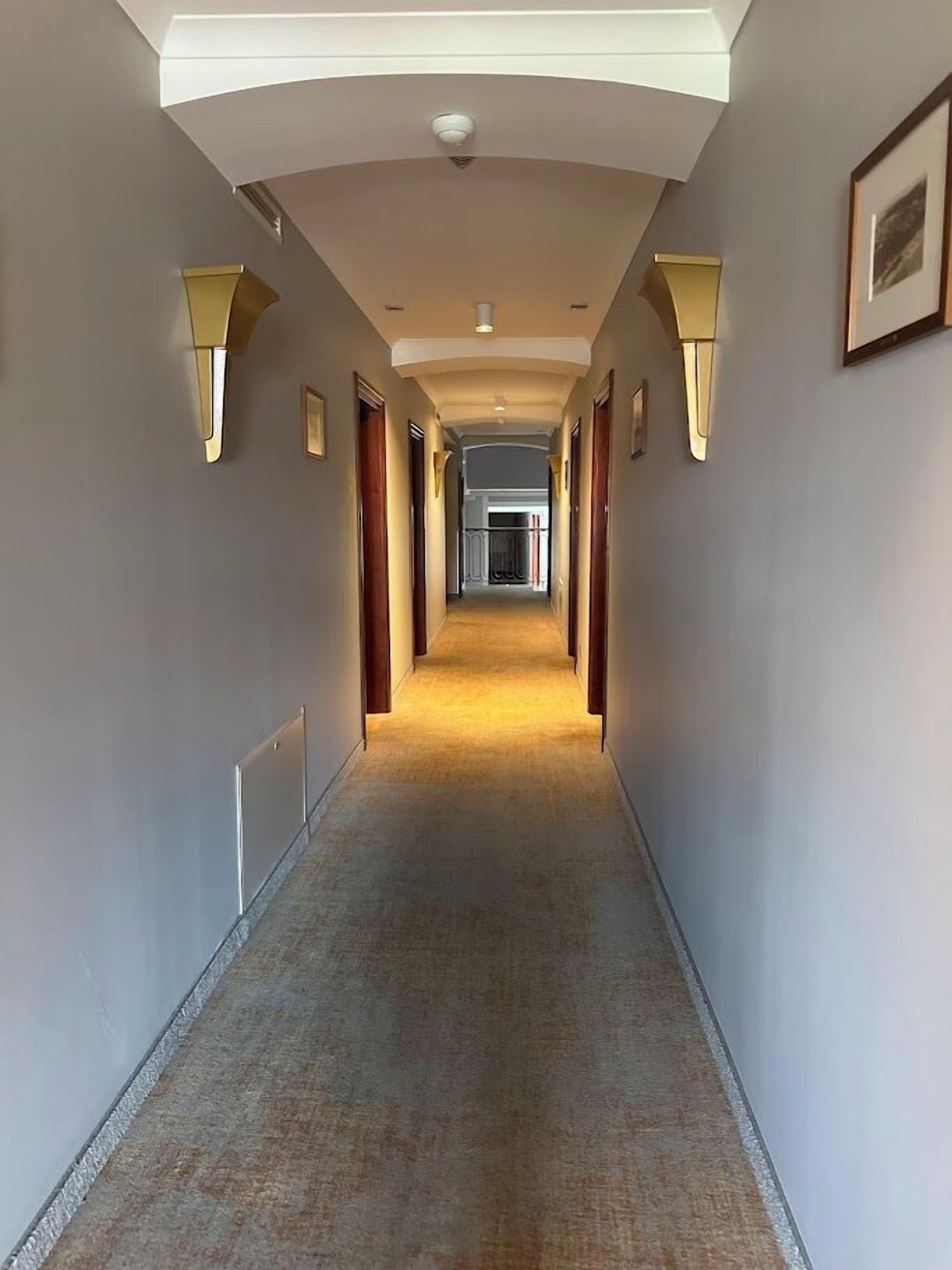A long, narrow hotel corridor with a carpeted floor, wall-mounted light fixtures, and framed pictures.