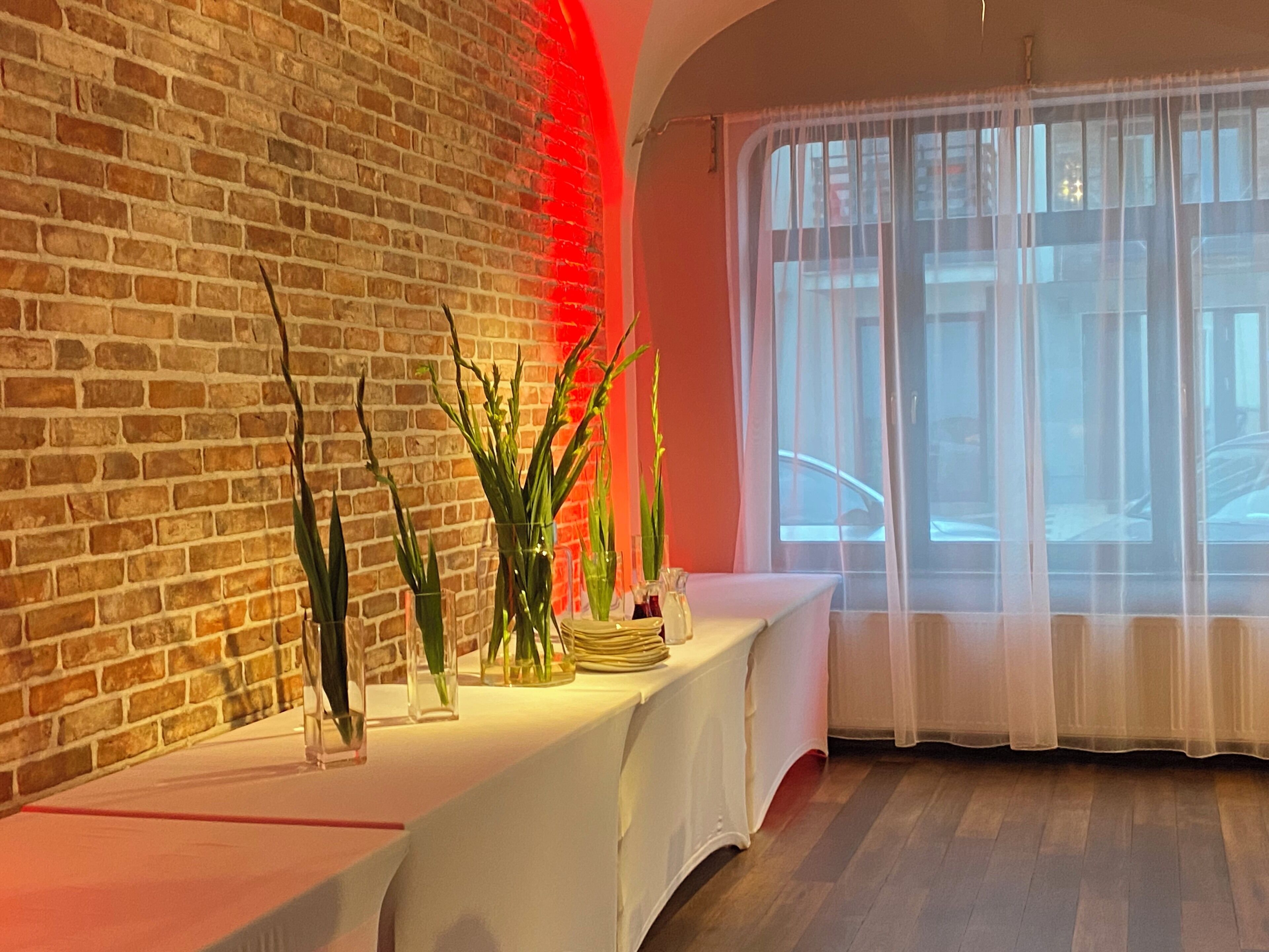 A room with a brick wall, a white table with a vase of plants, and a window with sheer curtains.