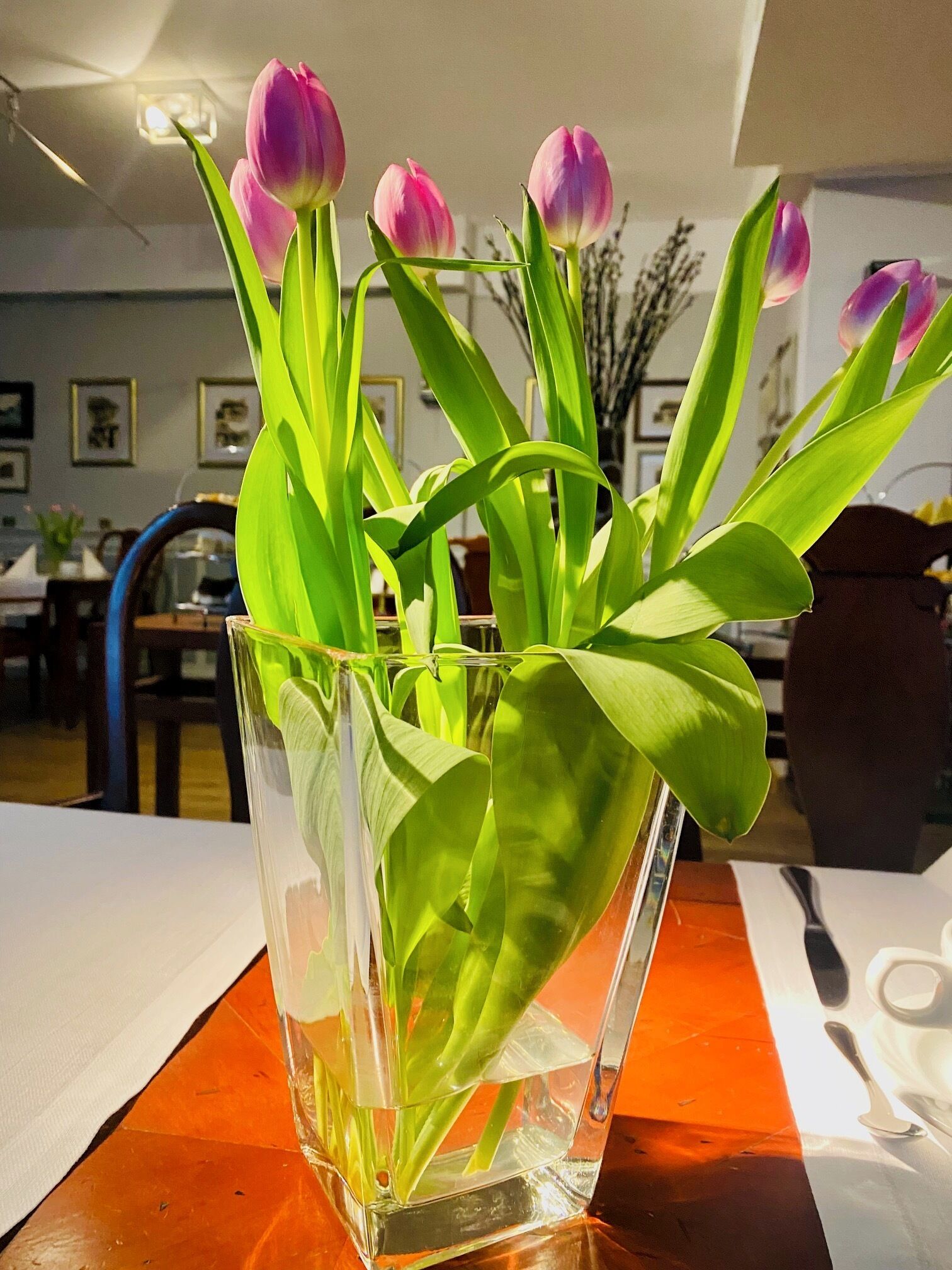 A clear glass vase with green leaves and pink tulips on a wooden table.