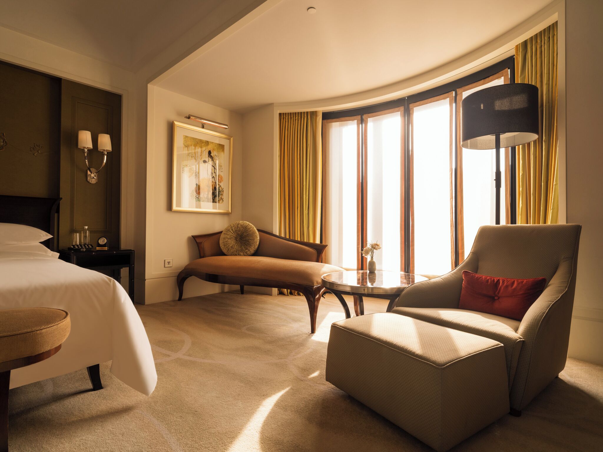 A hotel room with a bed, a sofa, a chair, a small table, and a large window with curtains.