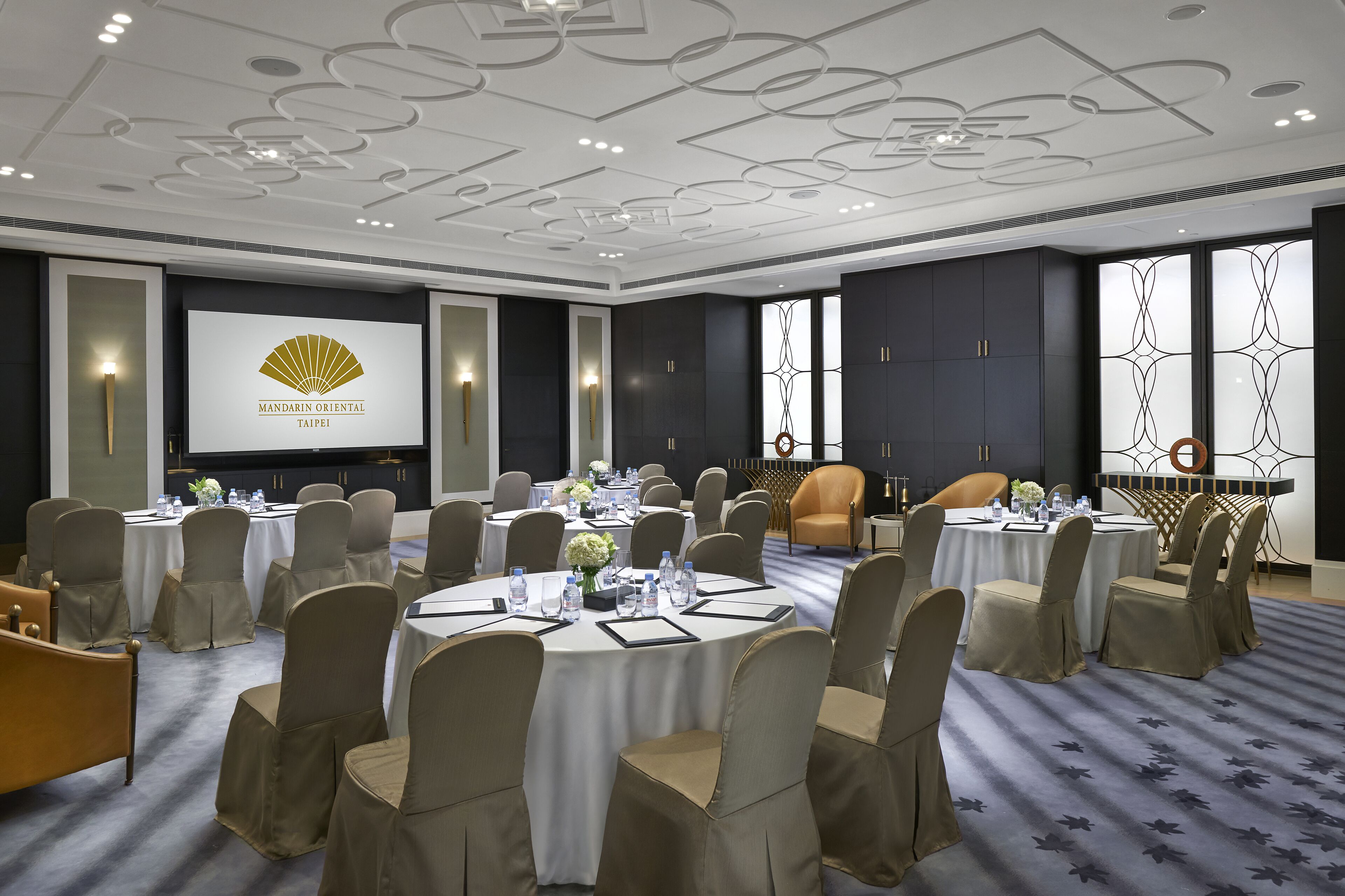 A conference room with round tables, beige chairs, and a large screen displaying the Mandarin Oriental Taipei logo.