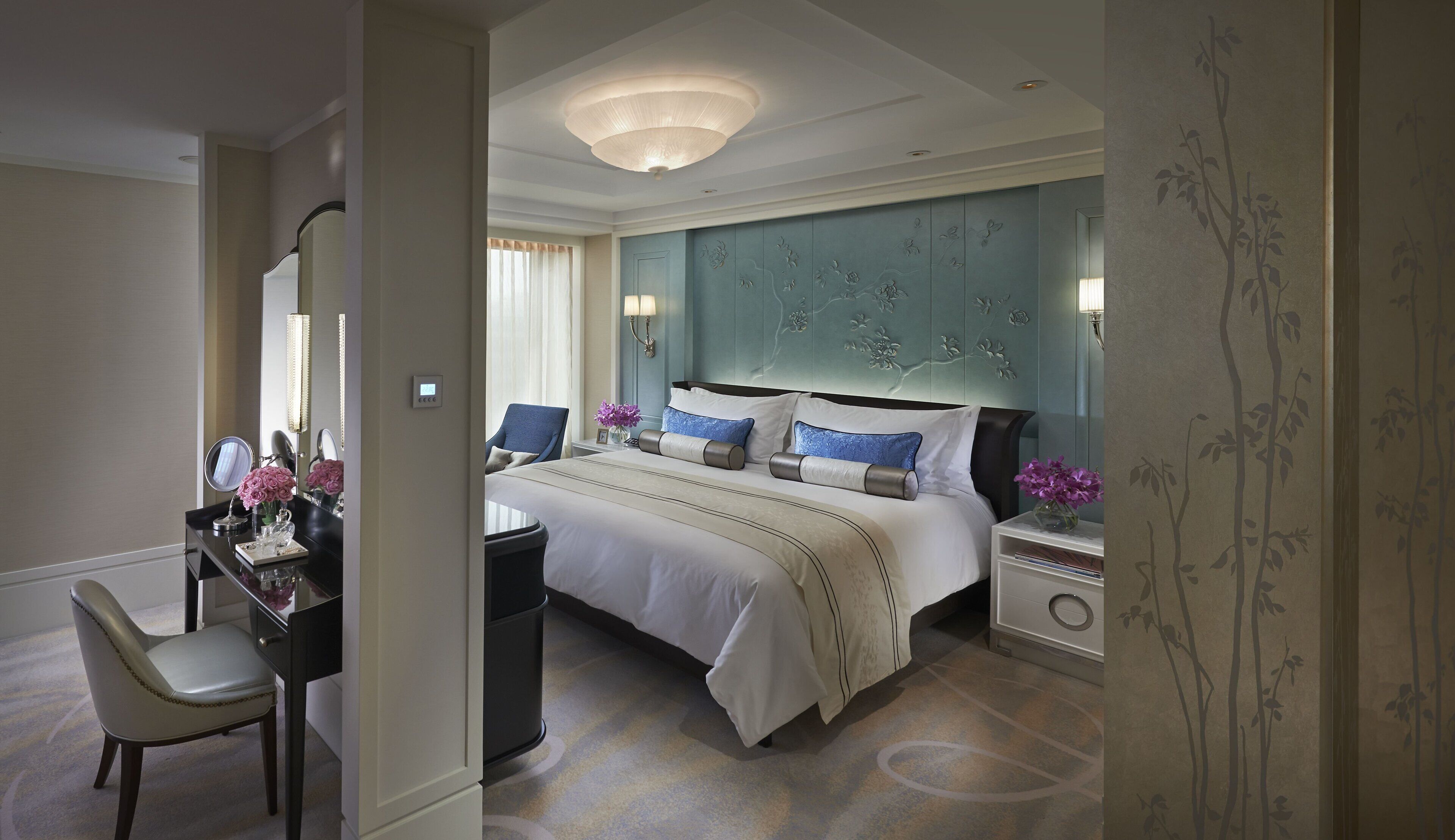 A hotel room with a large bed, a desk with a chair, a vanity with a mirror, and a nightstand with a lamp.
