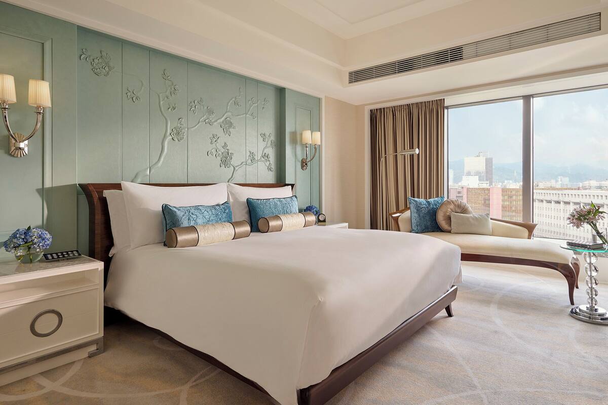 Ein Hotelzimmer mit einem großen Bett, einem Nachttisch, einem Schreibtisch mit Lampe, einem Sessel und Blick auf die Stadt durch große Fenster.
