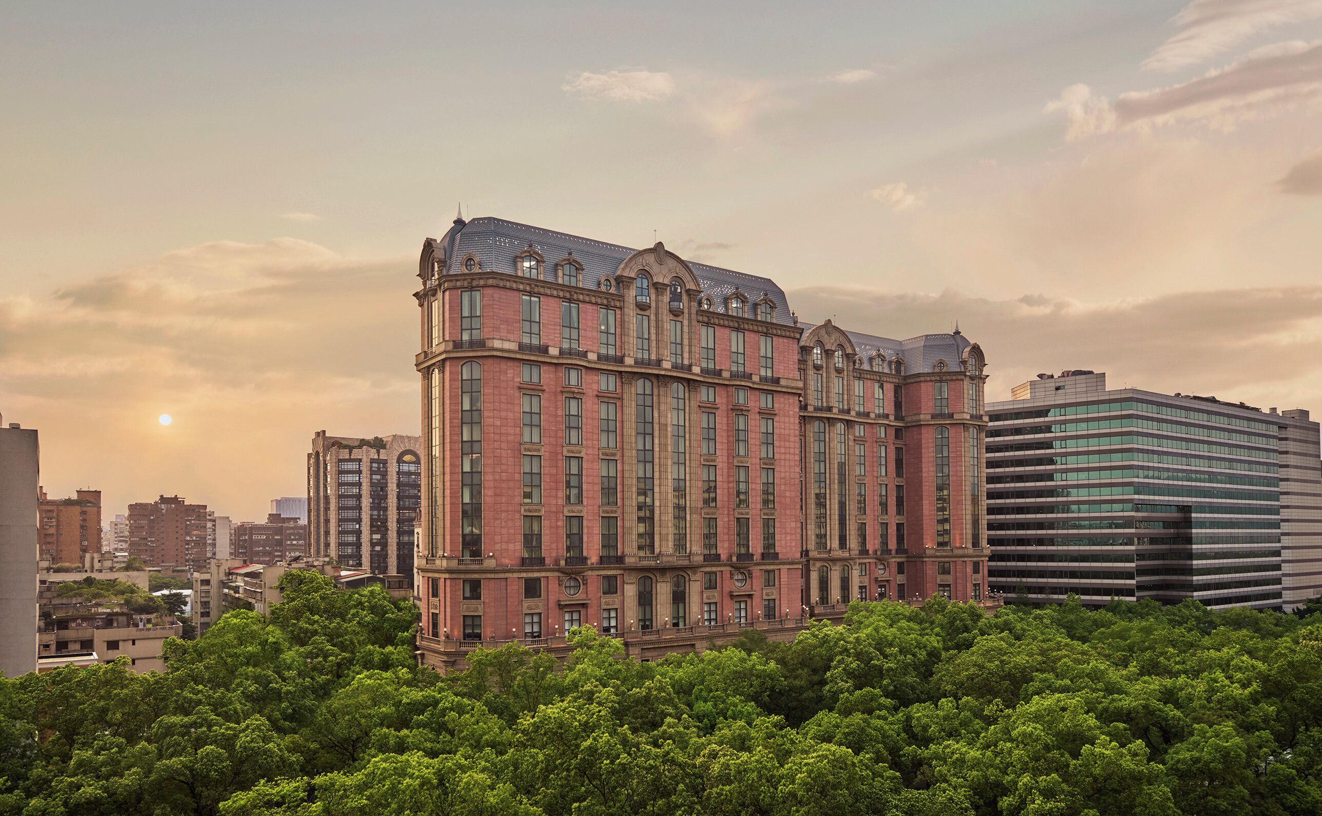 A large, historic building with multiple floors and a prominent corner turret, surrounded by dense green trees and modern high-rise buildings.