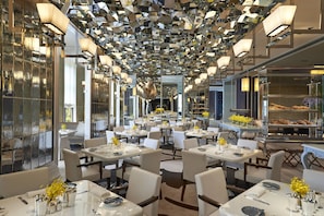 Um restaurante moderno com mesas e cadeiras brancas, e teto feito de painéis refletivos.