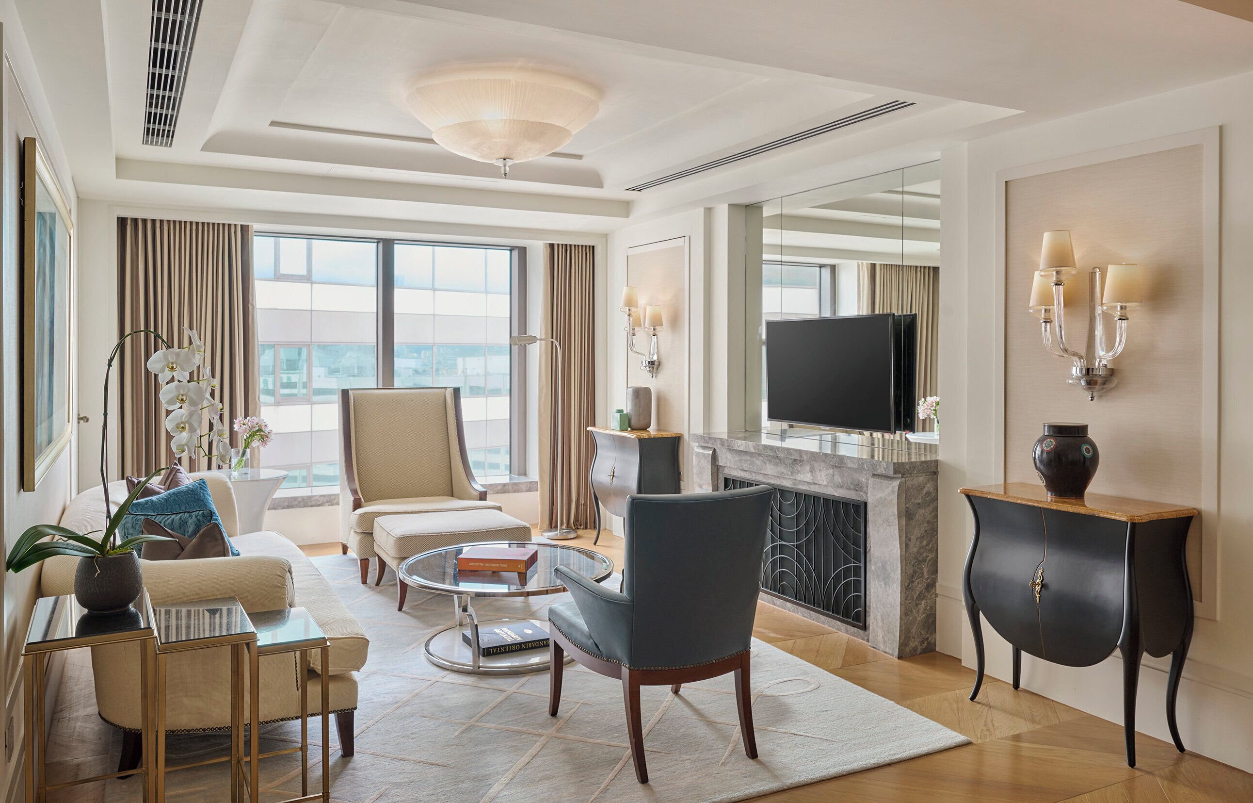 A modern hotel room with a sofa, armchairs, a coffee table, a TV, and a fireplace.