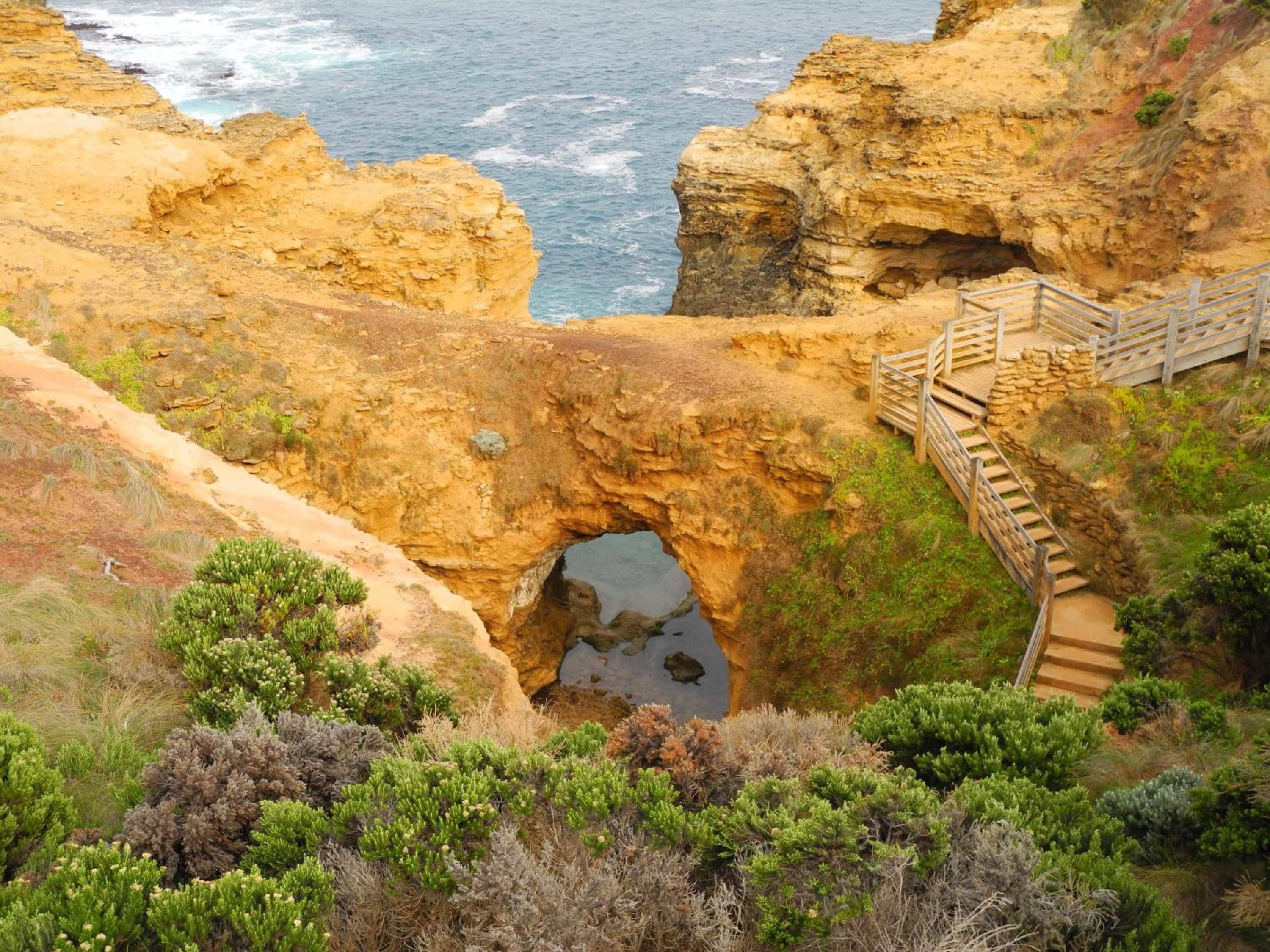 A coastal landscape with a natural rock formation, a wooden staircase leading to a viewing platform, and a clear view of the ocean.
