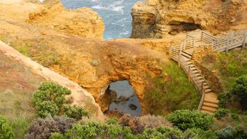 A coastal landscape with a natural rock formation, a wooden staircase leading to a viewing platform, and a clear view of the ocean.