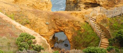 A coastal landscape with a natural rock formation, a wooden staircase leading to a viewing platform, and a clear view of the ocean.