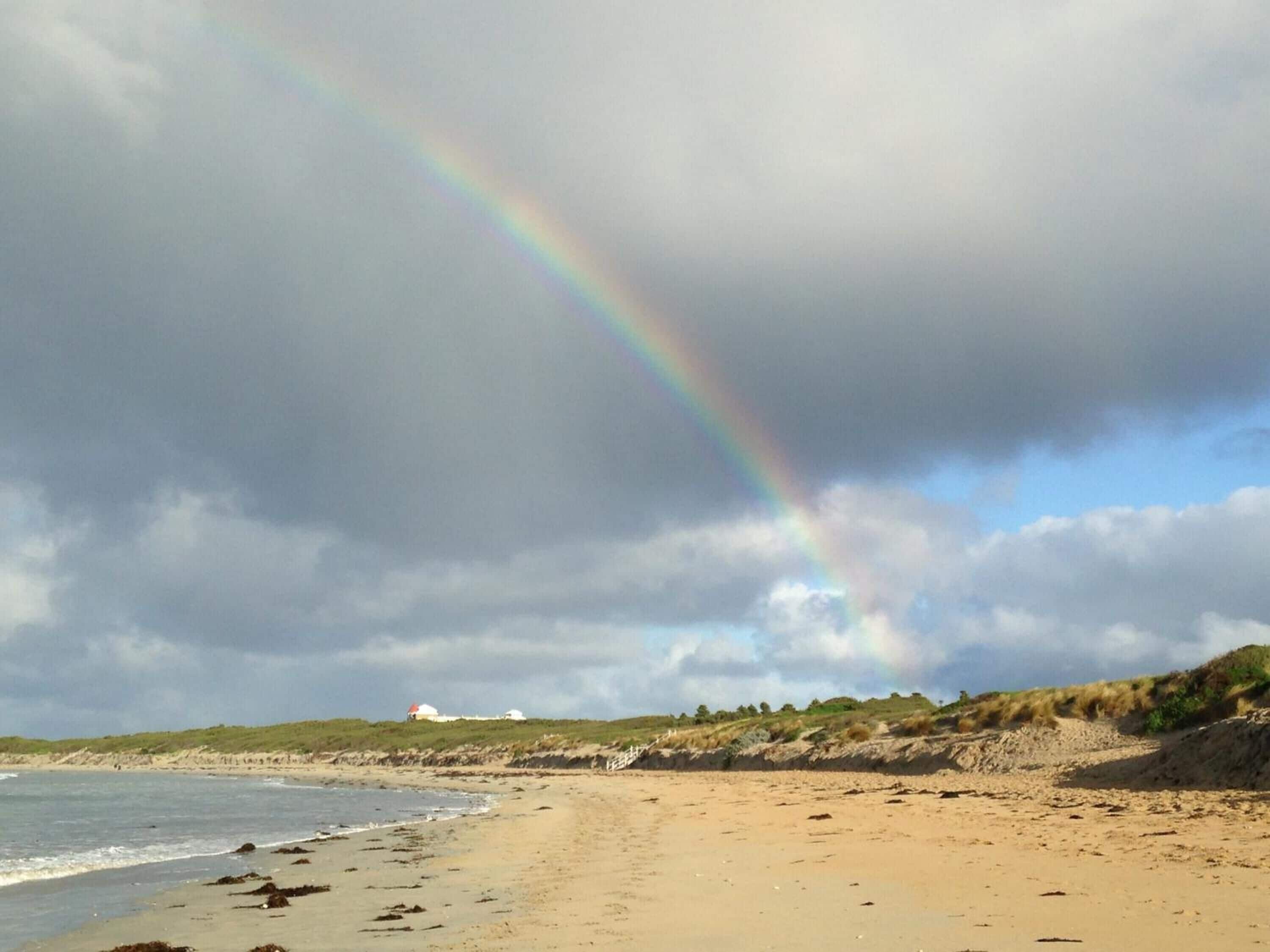 A beach with a rainbow, sandy shore, and dunes in the background.