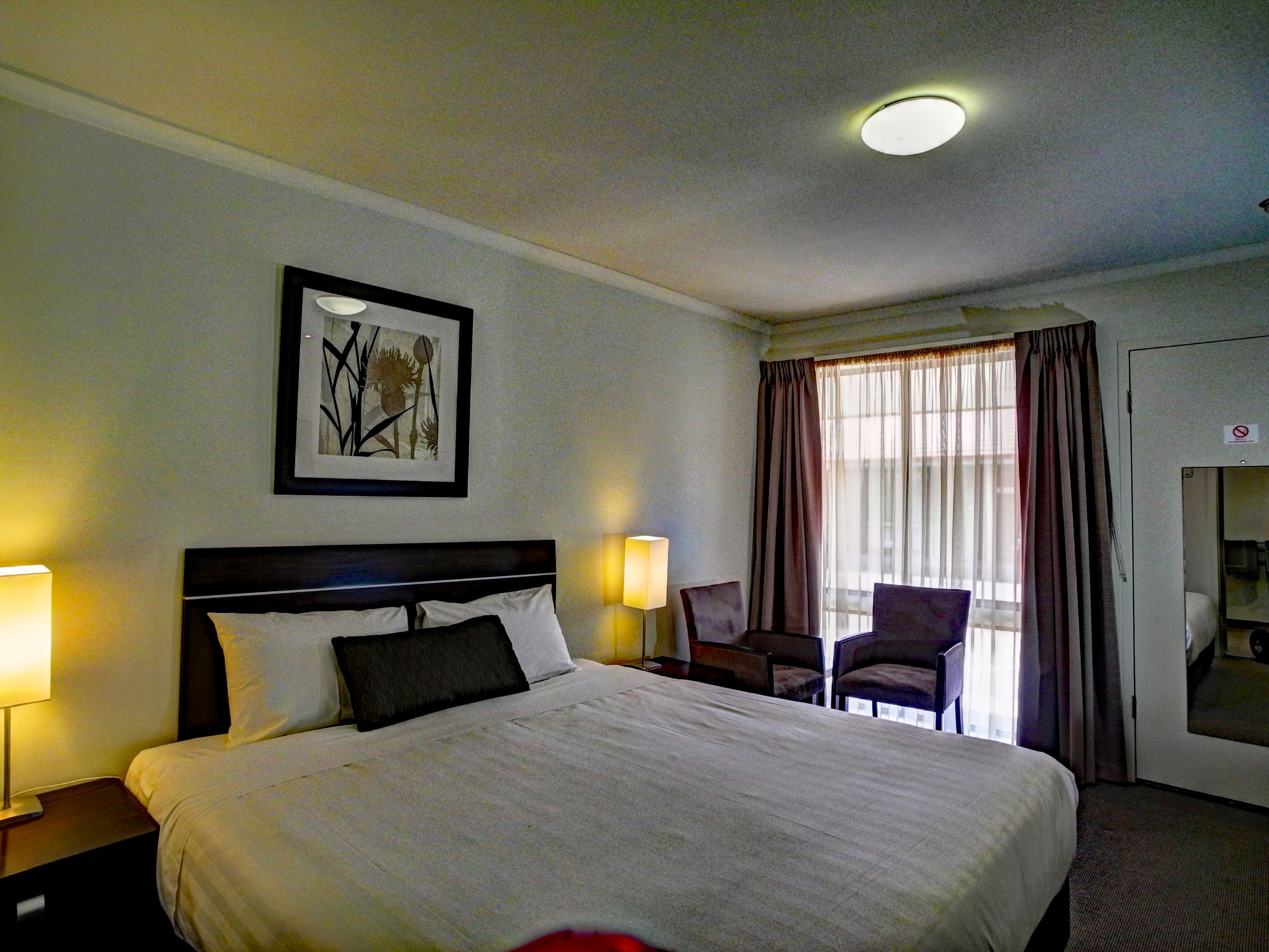 Une chambre d’hôtel avec un lit, deux lampes de chevet, une chaise, une fenêtre avec des rideaux et un tableau encadré au mur.