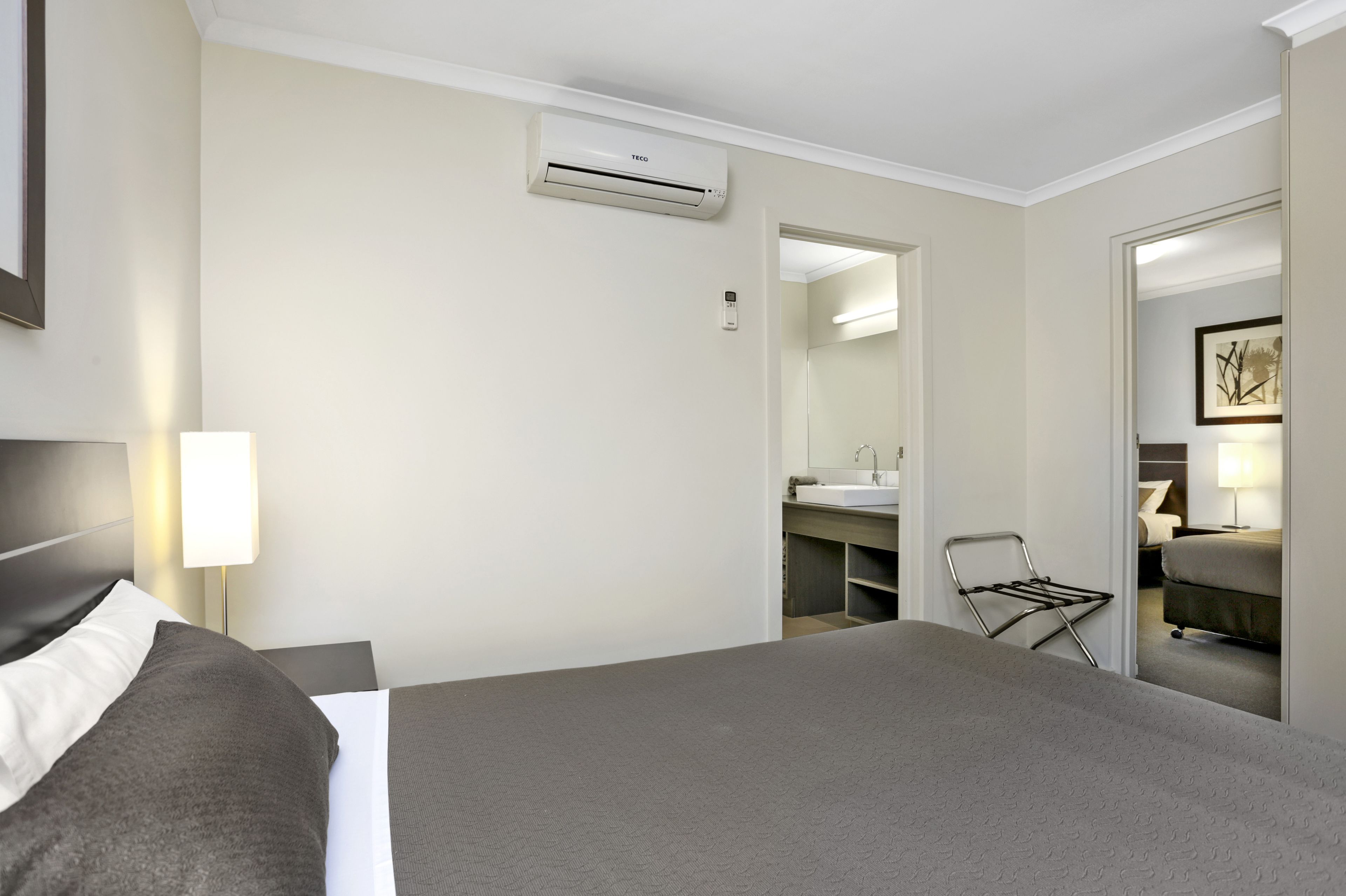 A hotel room with a bed, a bedside lamp, a desk with a chair, a mirror, and an air conditioning unit.
