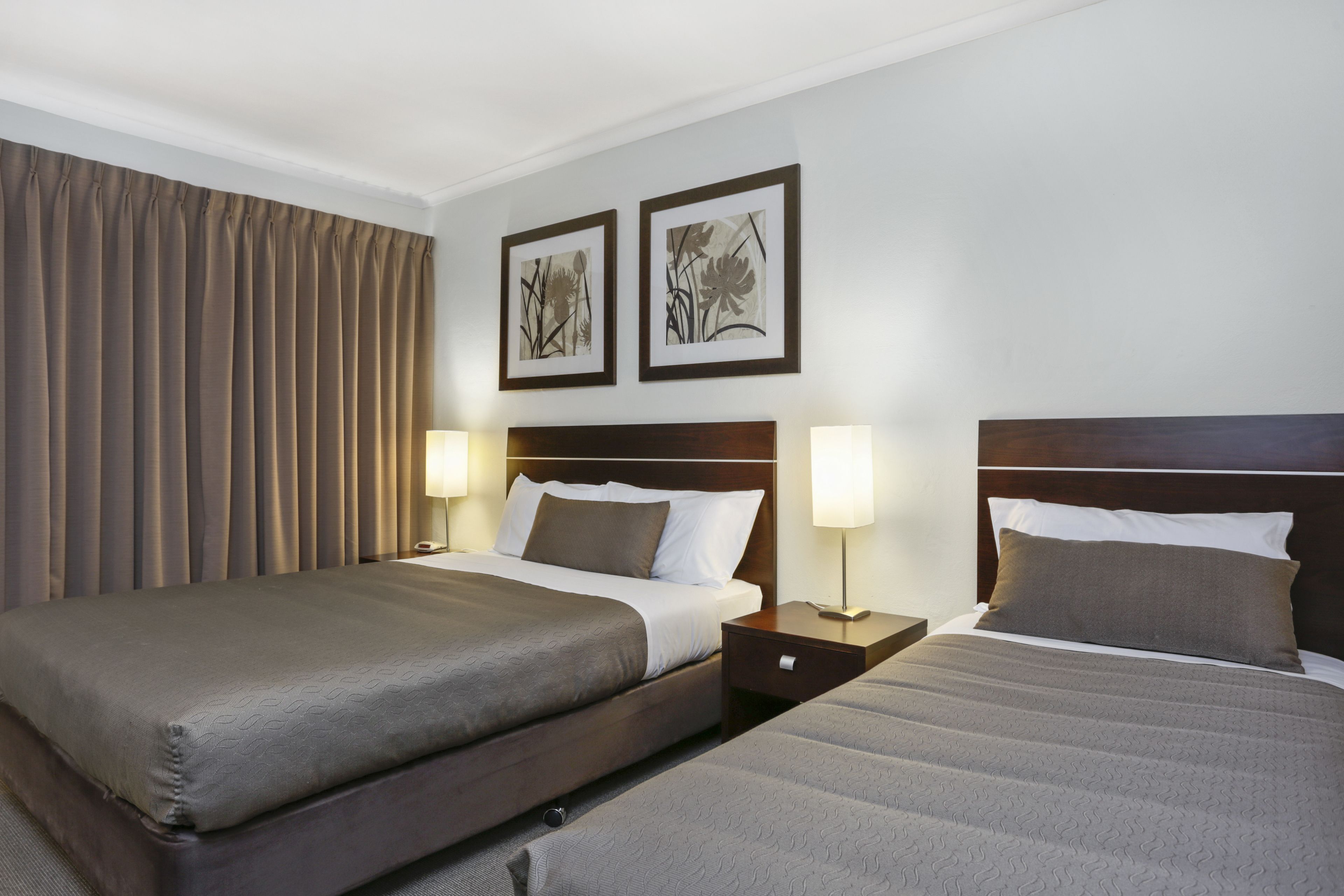 A hotel room with two beds, a wooden headboard, a nightstand with a lamp, and two framed pictures on the wall.