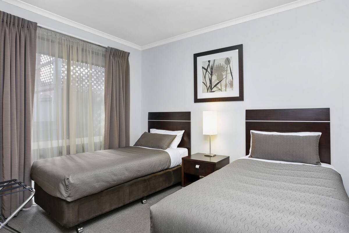 Habitación de hotel con dos camas, una mesita de noche, una lámpara, una ventana con cortinas y un cuadro en la pared.