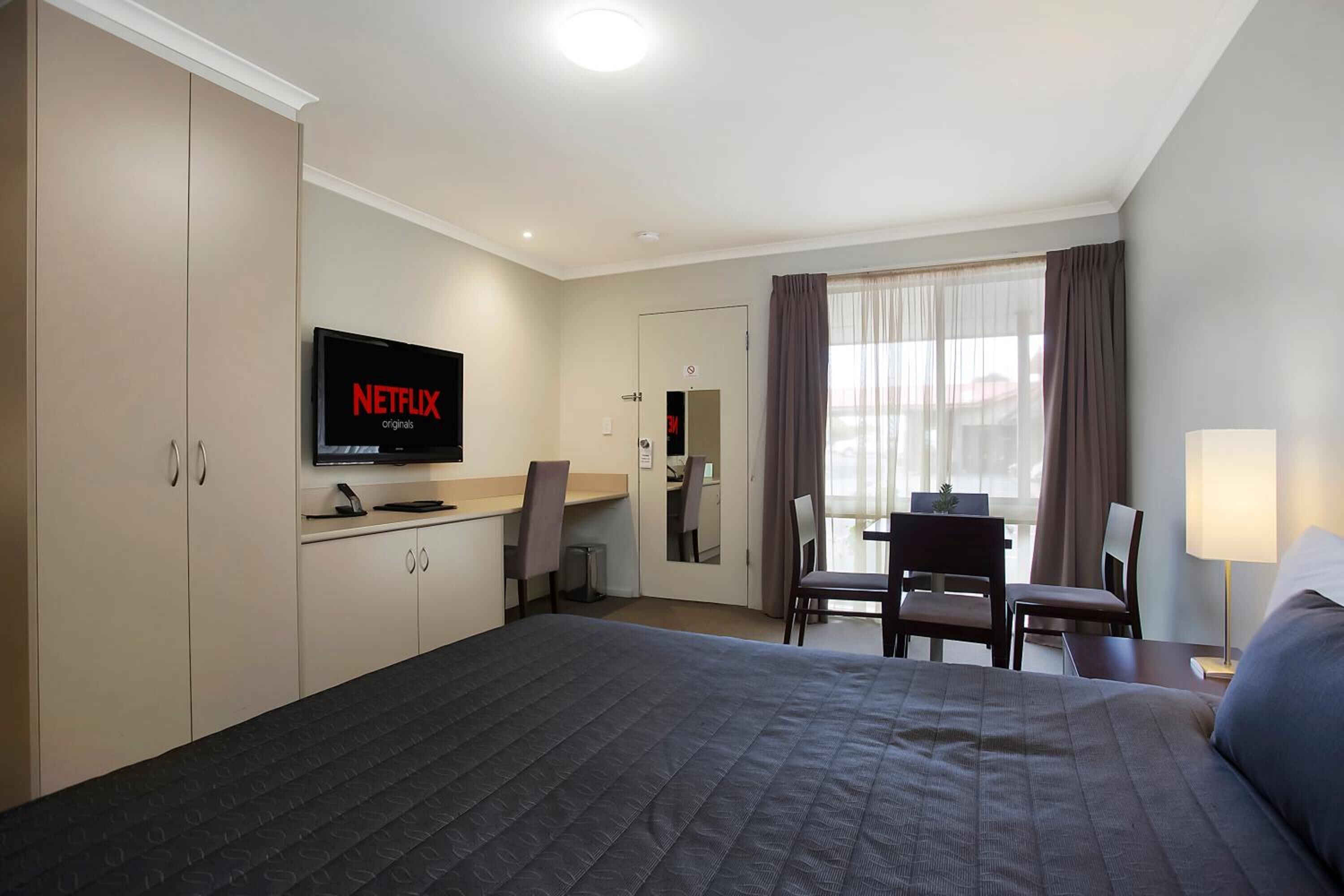 A hotel room with a bed, a desk with a chair, a television displaying 'NETFLIX originals', and a window with curtains.