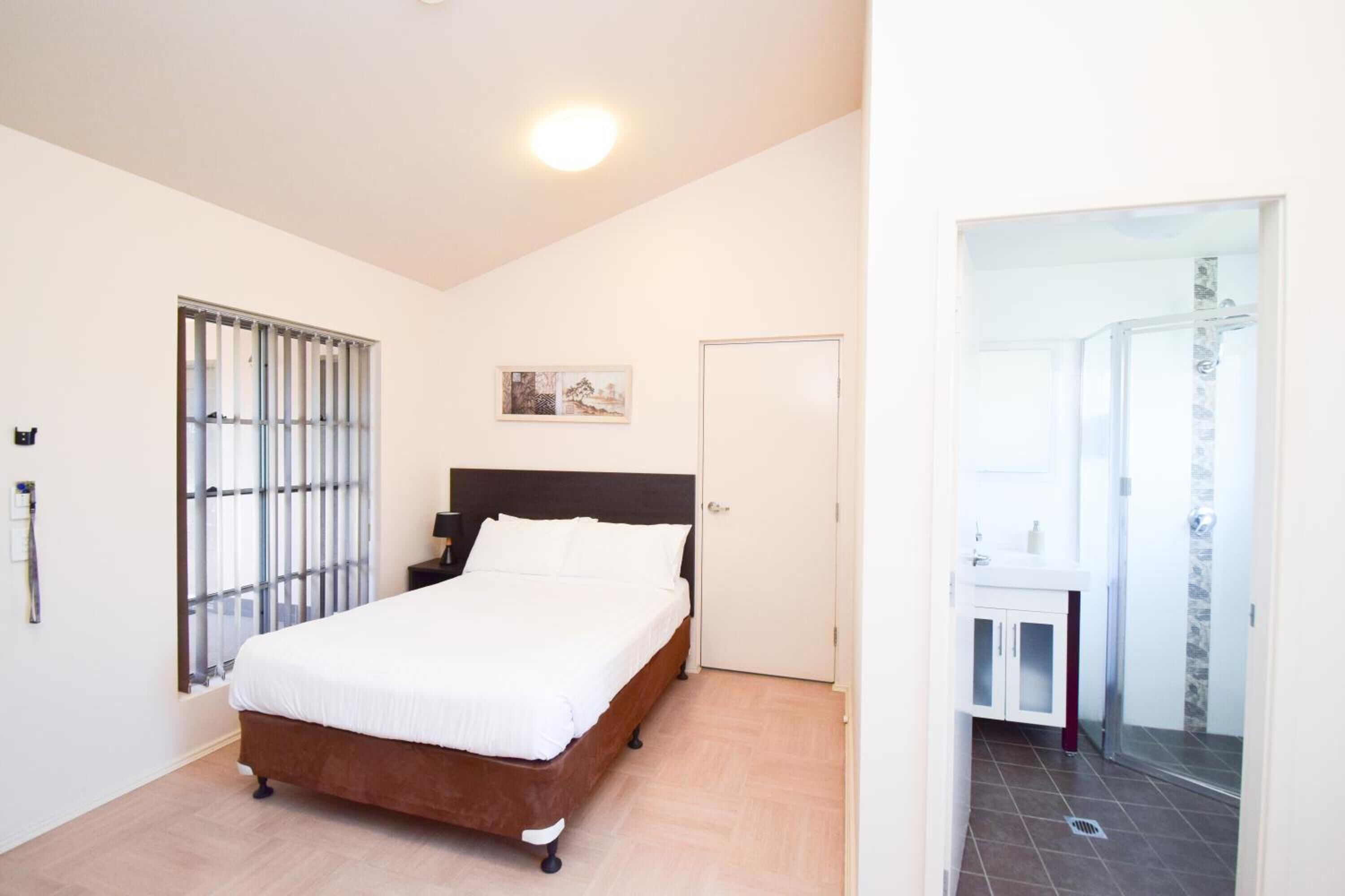 Un dormitorio con cama, ventana con persianas, puerta y baño visible a través de una puerta abierta.