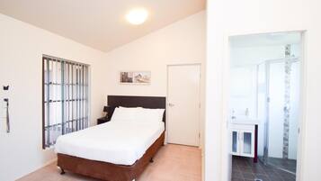 Un dormitorio con cama, ventana con persianas, puerta y baño visible a través de una puerta abierta.