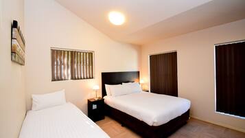 Quarto de hotel com duas camas, cabeceira, mesas de cabeceira com abajures, uma obra de arte na parede e uma janela com persianas.