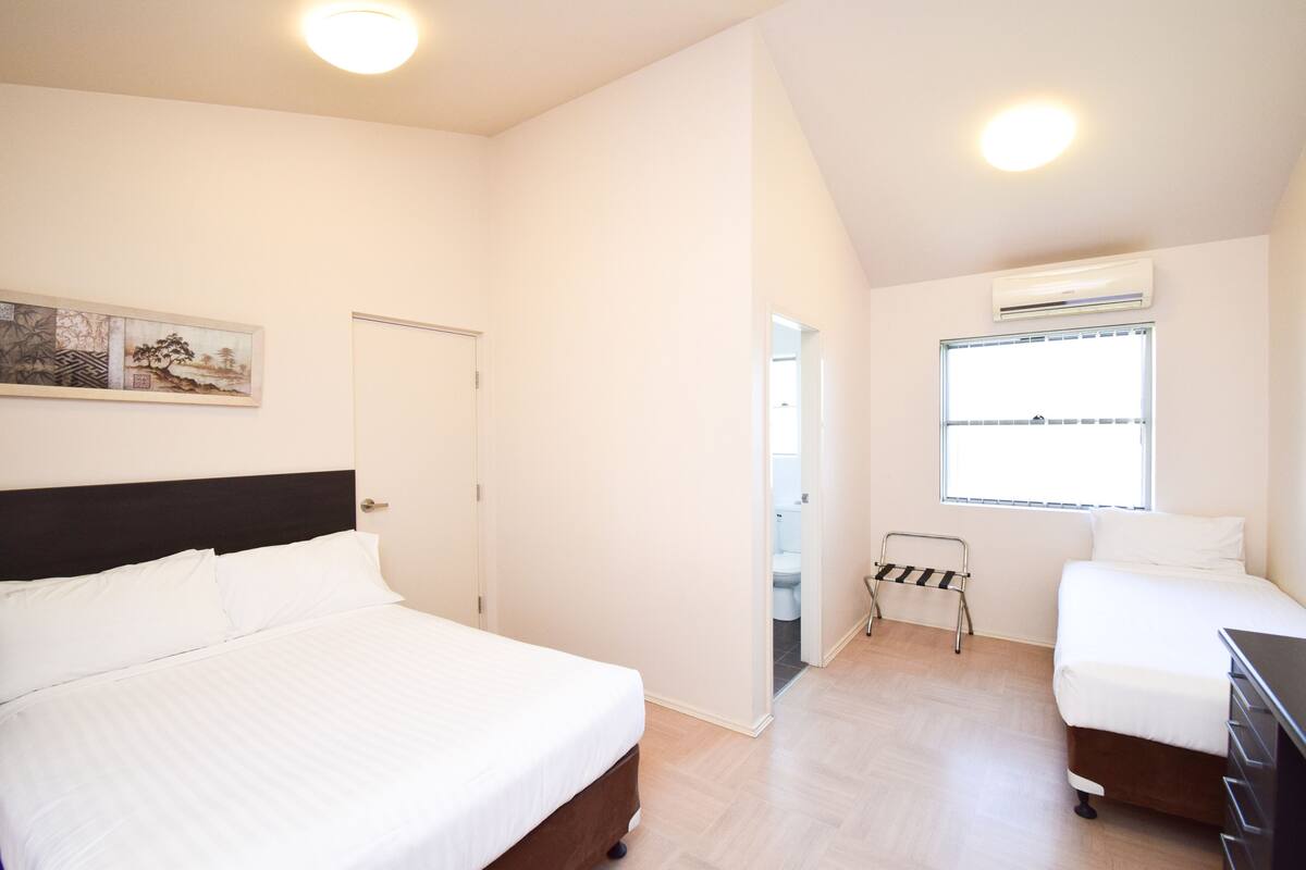 A bedroom with two beds, a wooden floor, a white wall, a painting, a door, and an air conditioning unit.