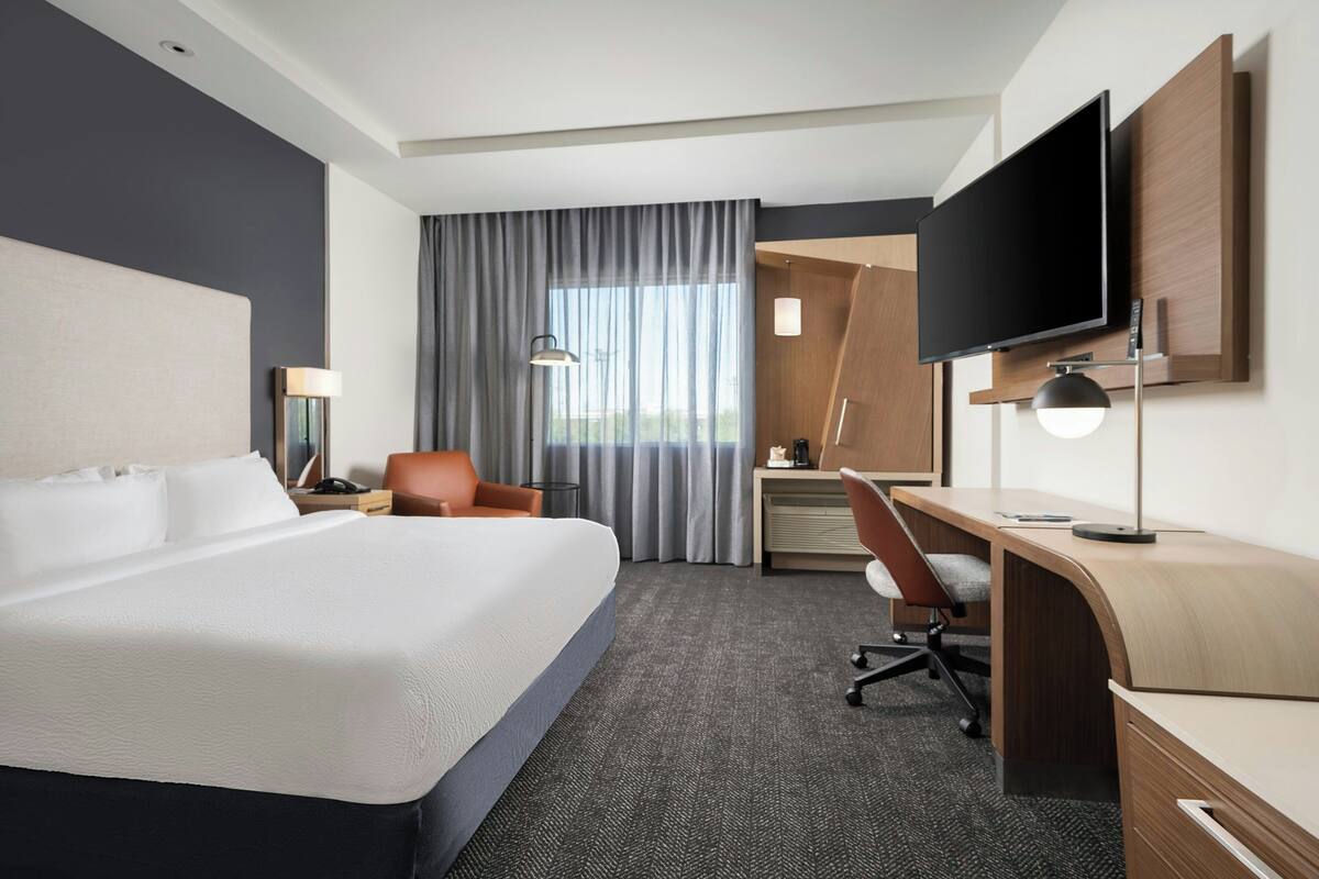 Habitación de hotel con cama, escritorio, silla, televisor y cortinas.