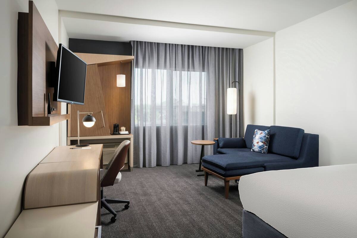 Habitación de hotel con cama, escritorio, silla, sofá, televisión, lámpara y ventana con cortinas.