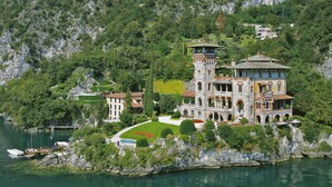 Un gran edificio parecido a un castillo, de varios pisos y con una torre, ubicado a orillas de un lago, con abundante vegetación y acantilados rocosos al fondo.