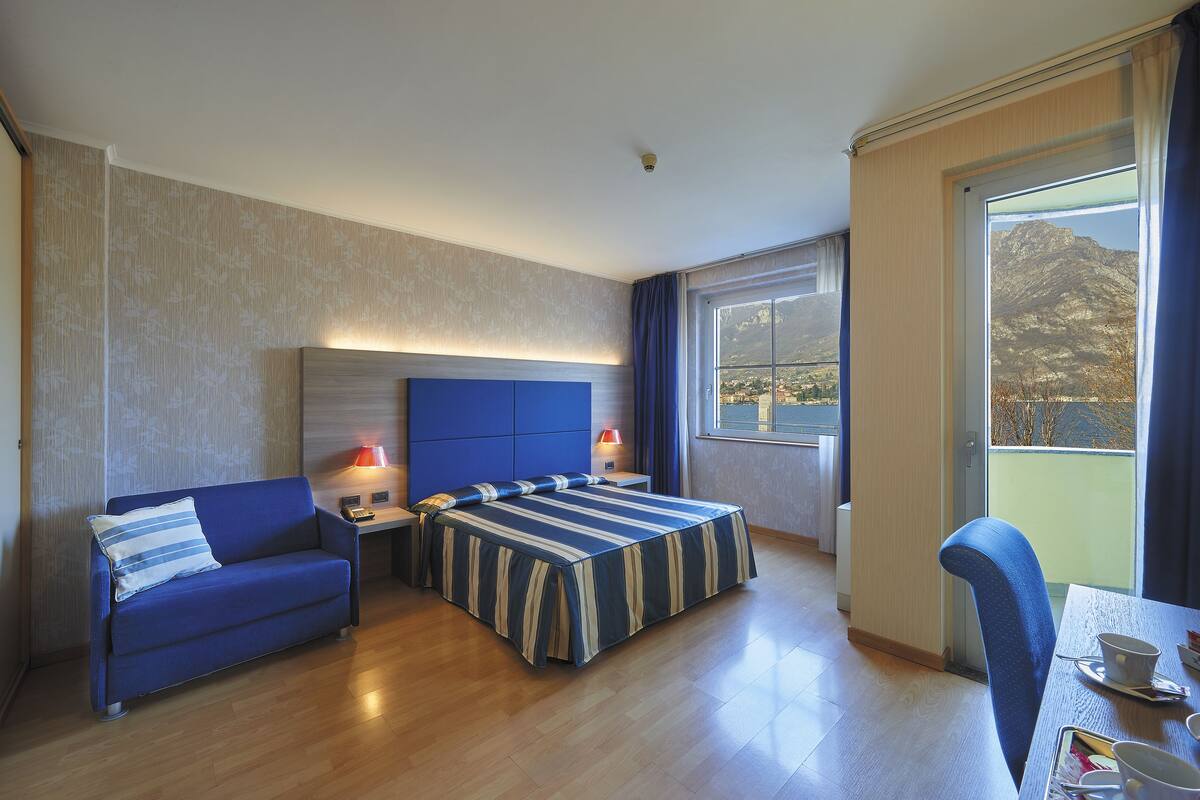 A hotel room with a bed, a sofa, a dining table, and a view of mountains through the window.
