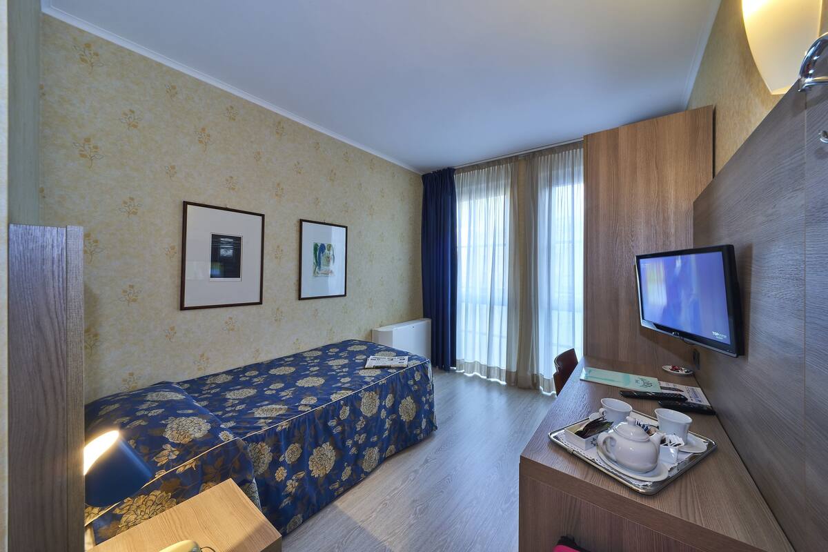 A hotel room with a single bed, a desk with a tray of tea and cups, a television, and two framed pictures on the wall.