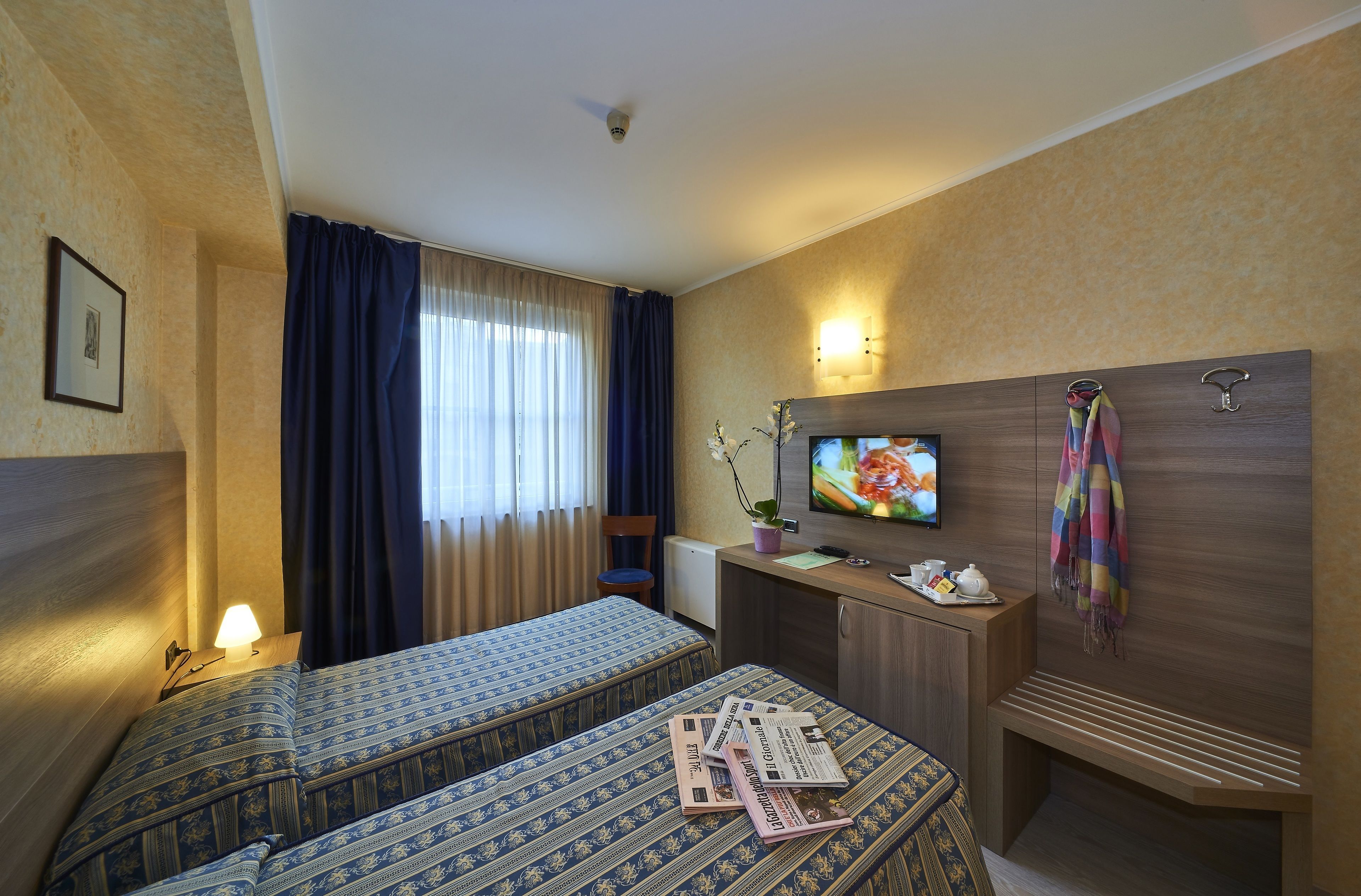 Camera d'albergo con due letti, una scrivania con televisore, una panca e un porta asciugamani a parete.