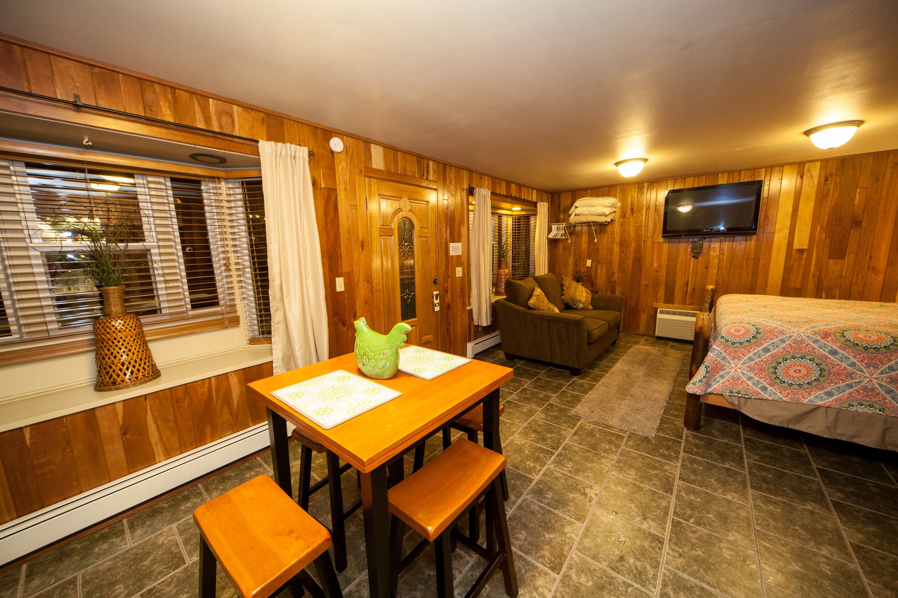 Una habitación acogedora con paredes de madera, una cama con colcha estampada, un televisor, una mesa de comedor con bancos y una ventana con persianas y cortinas.