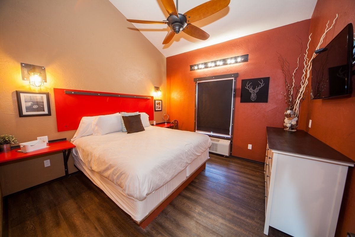 A hotel room with a bed, a red headboard, a ceiling fan, a bedside table, a nightstand, a dresser, a mirror, and wall-mounted lights.