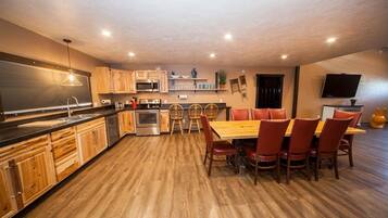 A modern kitchen with wooden cabinets, a dining area with red chairs, and a flat-screen TV mounted on the wall.
