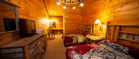 A log cabin room with a wooden ceiling, a bed with a tiger print blanket, a desk with a lamp, and a television on a dresser.