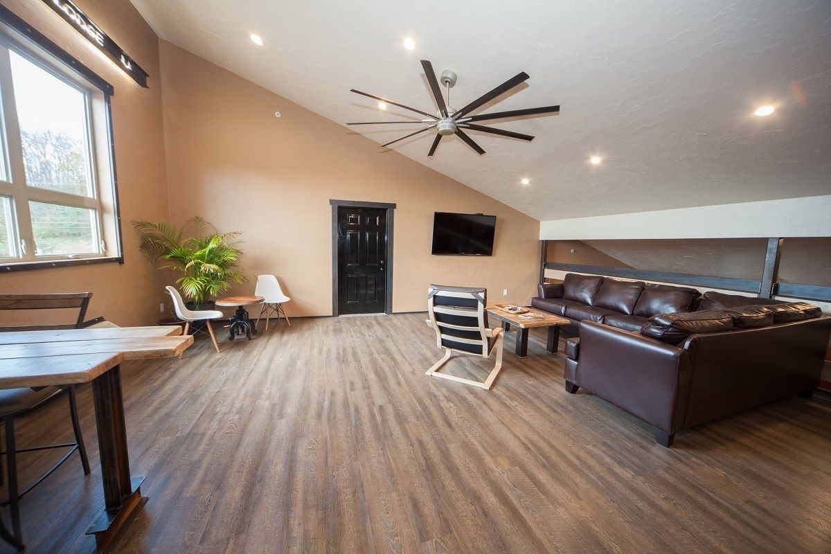 A spacious living area with a high ceiling, a ceiling fan, a brown leather sofa, a dining table, and a television mounted on the wall.