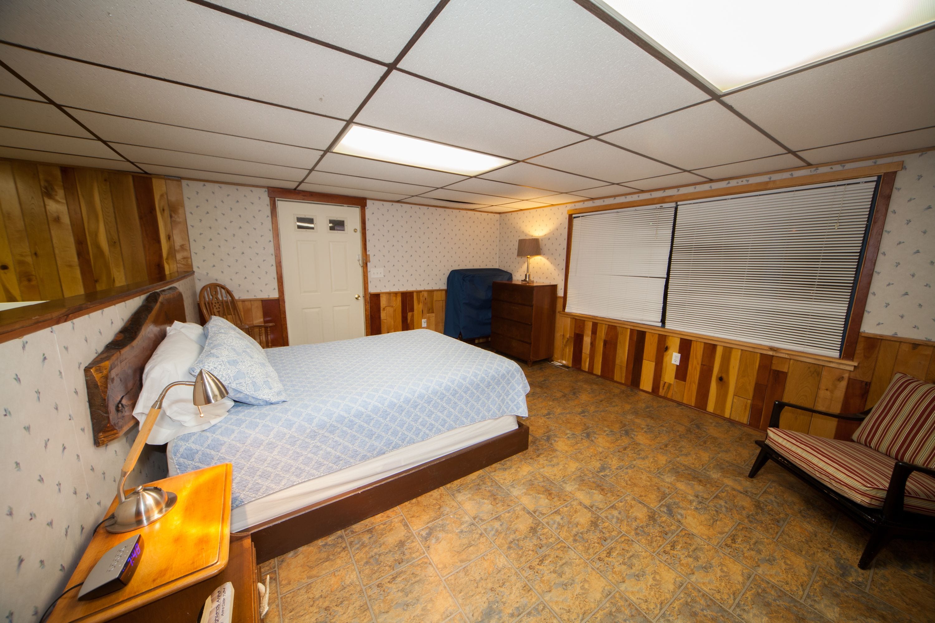Quarto com cama, uma cadeira, escrivaninha com luminária, janela com persianas e porta.