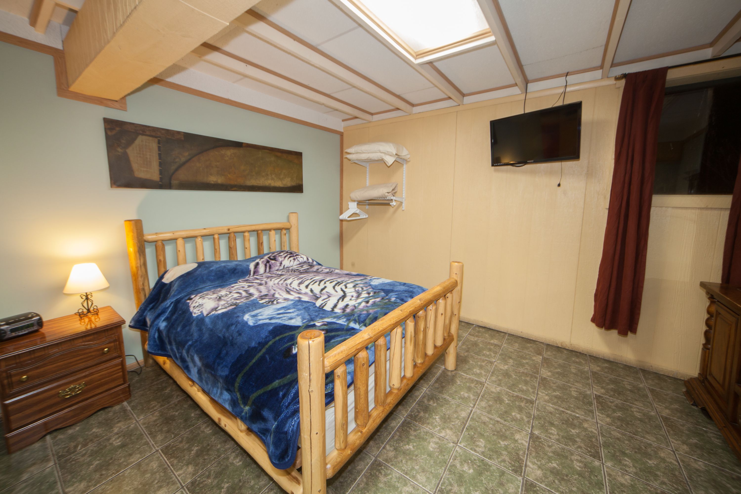 A bedroom with a wooden bed, a nightstand with a lamp, a wall-mounted TV, and a painting on the wall.