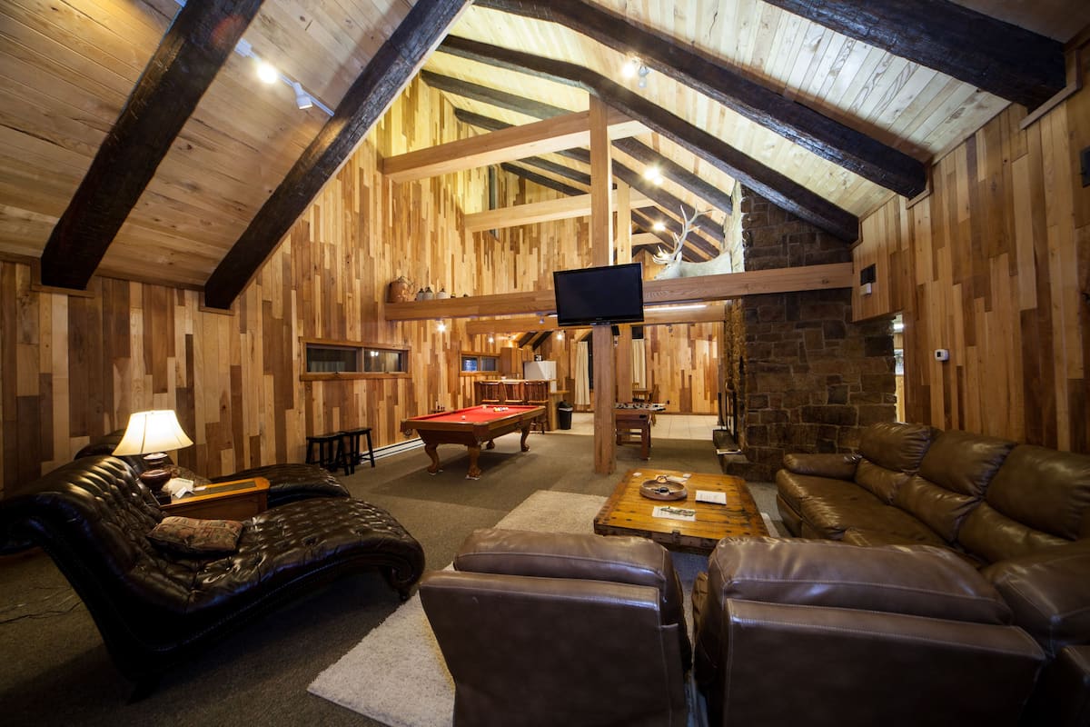 A cozy indoor space with wooden walls and a high ceiling, featuring a pool table, leather sofas, and a television mounted on the wall.