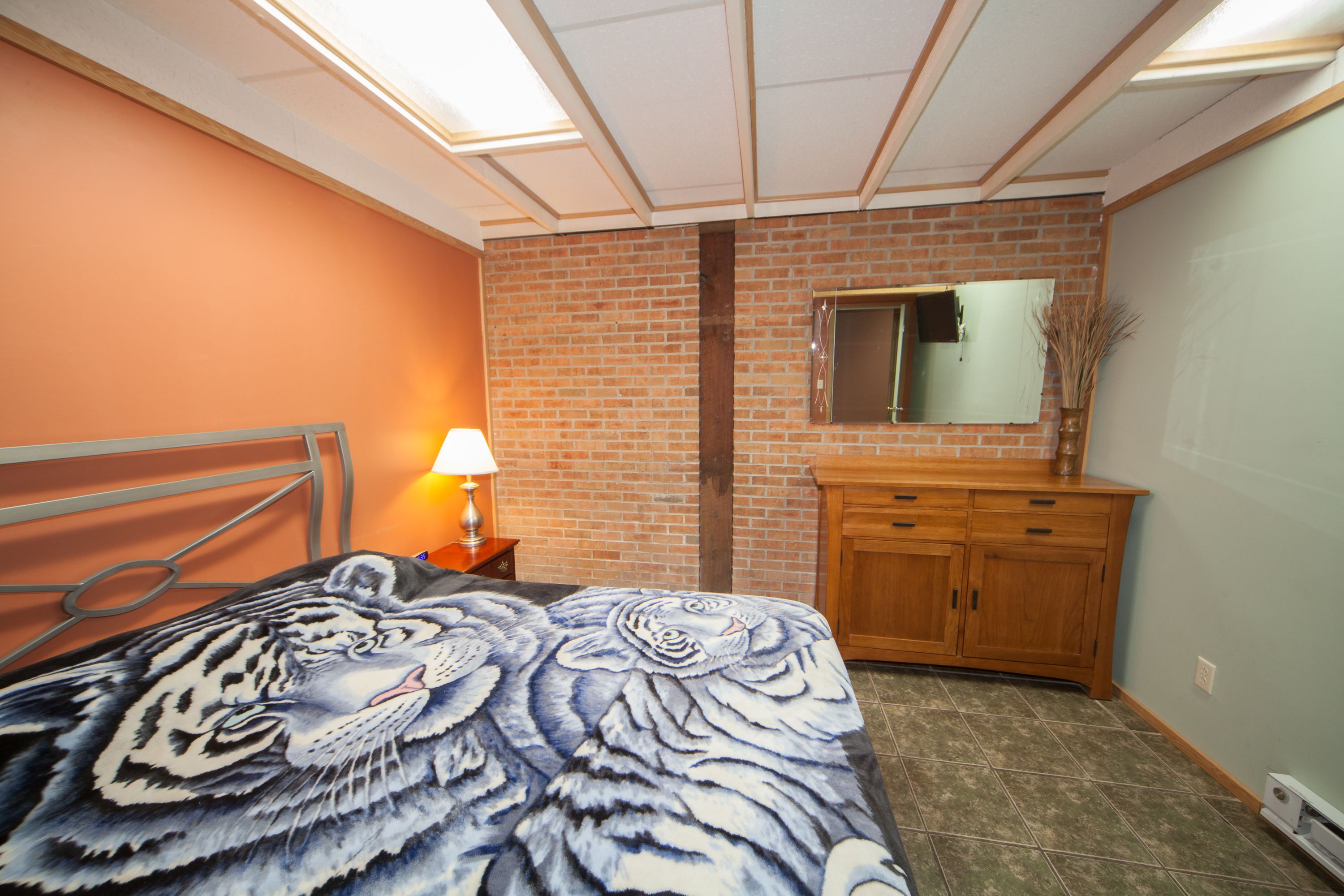 A bedroom with a bed, a nightstand, a wooden dresser, a mirror, and a brick wall.