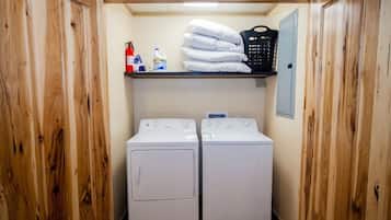 A laundry room with two white washing machines, a shelf with towels and cleaning supplies, and a fire extinguisher.