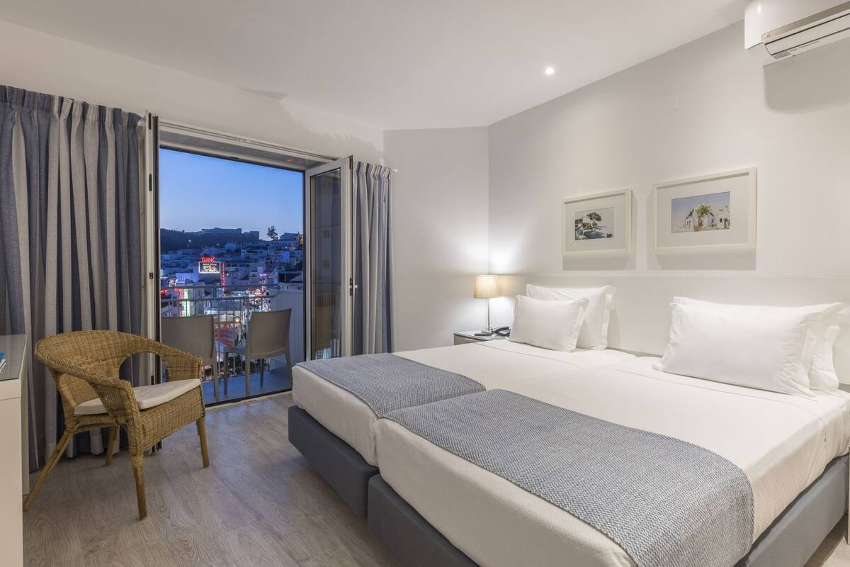 Ett hotellrum med en stor säng, en stol, ett skrivbord och en balkong med utsikt över staden.