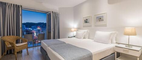 A hotel room with a bed, a chair, a nightstand, and a view of the city at dusk.