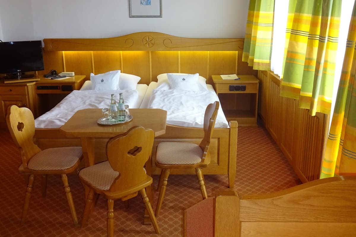 Une chambre d’hôtel avec une tête de lit en bois, deux lits avec du linge de lit blanc, une table en bois avec des chaises, une télévision et un tableau encadré au mur.